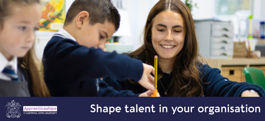 A Teacher leans down with her student who is placing his pencil into a pencil grip. The teacher is smiling and supporting the student.
